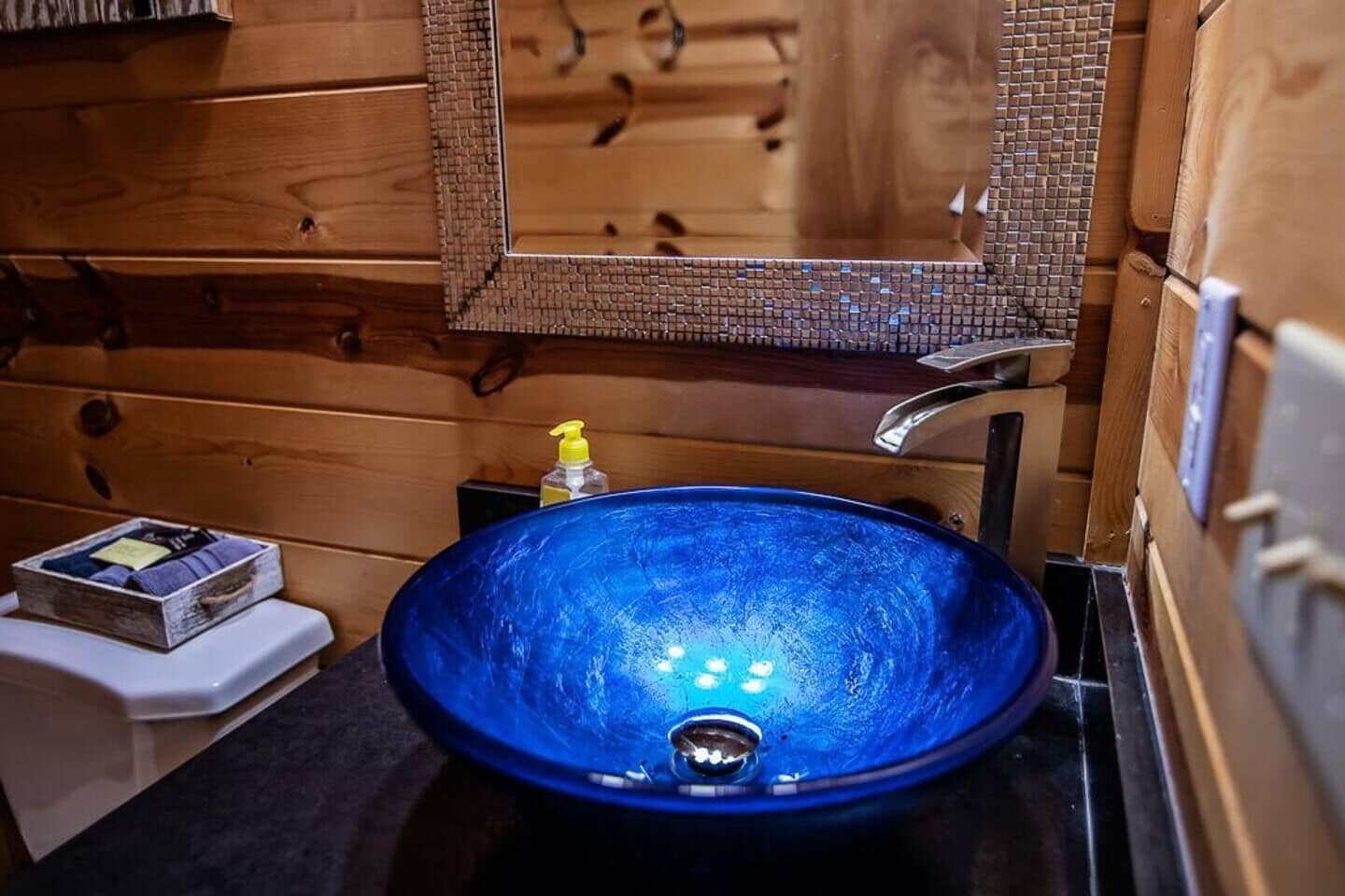 Un baño con un lavabo de vidrio azul, paredes de madera, un espejo y un pequeño estante con artículos de tocador.