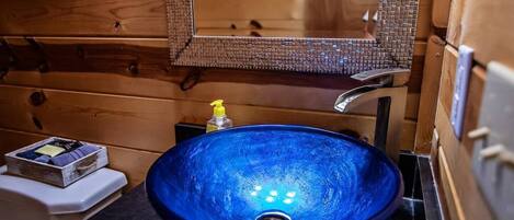 Un baño con un lavabo de vidrio azul, paredes de madera, un espejo y un pequeño estante con artículos de tocador.