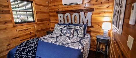 Ein Schlafzimmer einer Holzhütte mit einem Bett, einem Nachttisch, einer Klimaanlage und einem Fenster mit Jalousien.