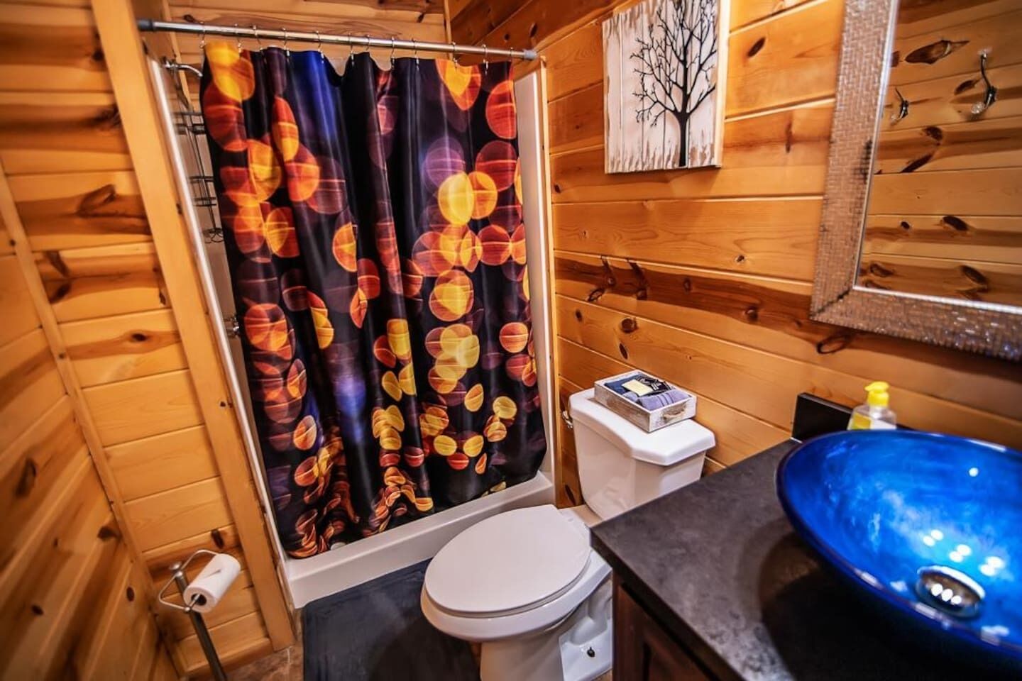Baño de cabaña de madera con cortina de ducha de diseño circular, un inodoro blanco, un lavamanos con recipiente azul y un espejo en la pared.