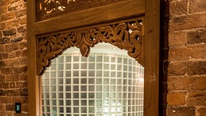 A mirror with ornate wooden carvings reflecting a room with a brick wall, a cushioned bench, and a window with a grid pattern. - Infinity Apartments Victoria Street (Liverpool)
