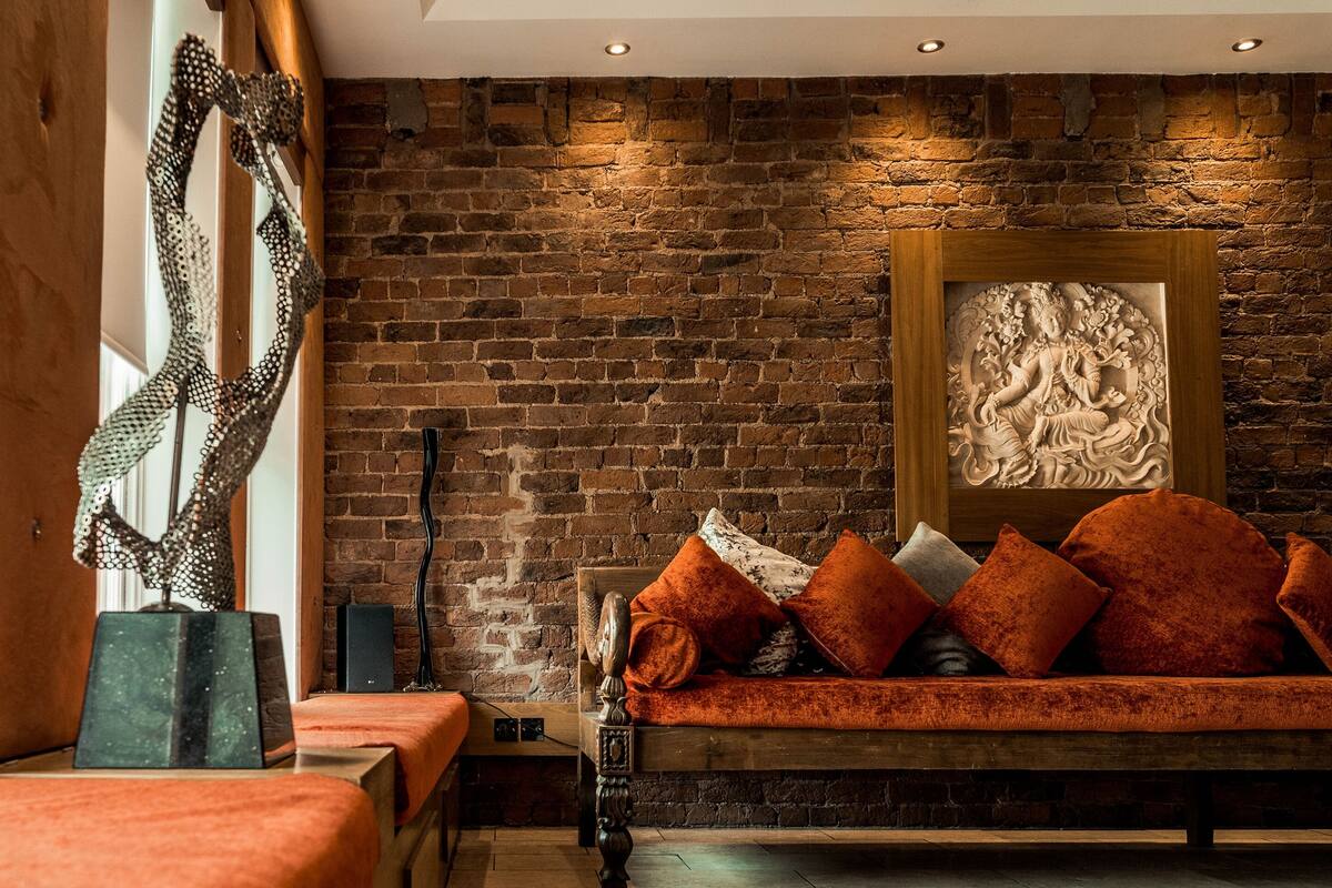 Ein Zimmer mit einer Ziegelwand, einer hölzernen Bank mit orangefarbenen Kissen und einer gerahmten Skulptur.