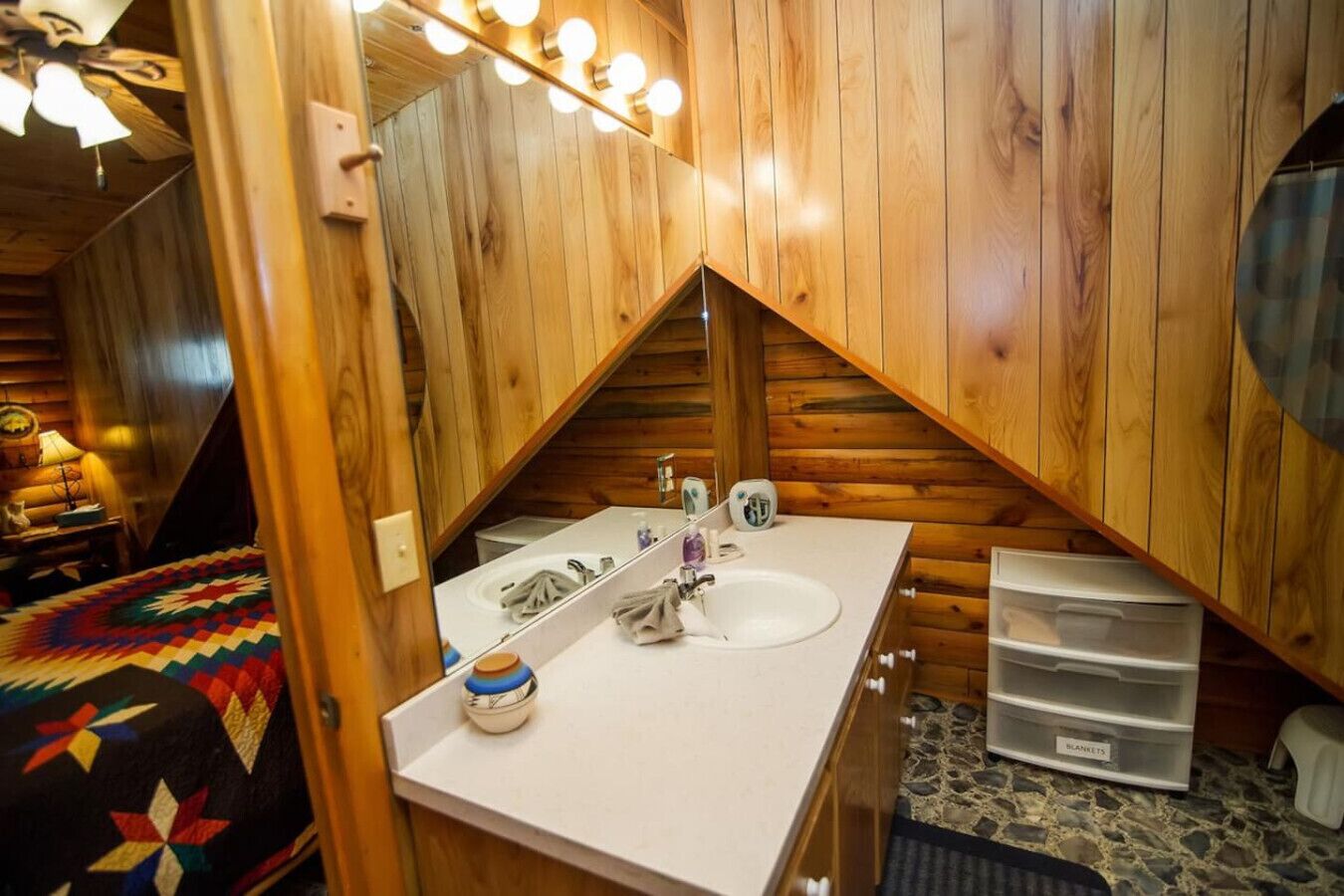 Ein gemütliches Badezimmer im Blockhausstil mit Holzvertäfelung, einem weißen Waschbecken, einem Spiegel mit Beleuchtung und einer gesteppten Tagesdecke.