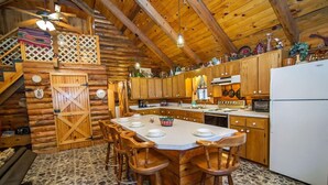 Interior rústico de una cabaña de troncos con una mesa de comedor central rodeada de sillas de madera, una zona de cocina con armarios de madera y un piso de baldosas de piedra.