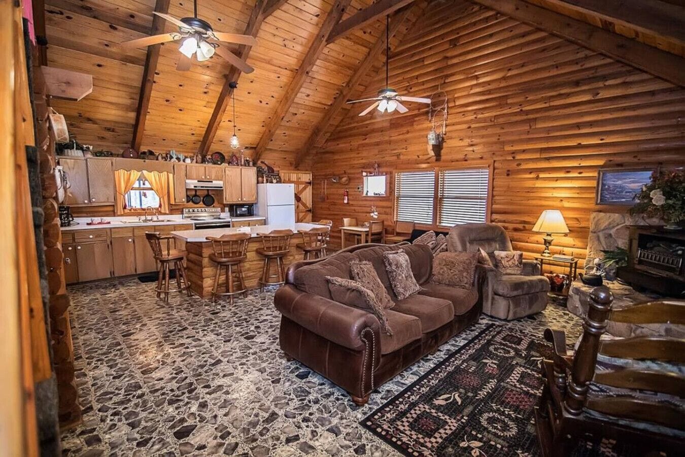 A log cabin interior with a stone-tiled floor, a brown leather sofa, a dining area with wooden chairs, a kitchen with wooden cabinets, and a fireplace.