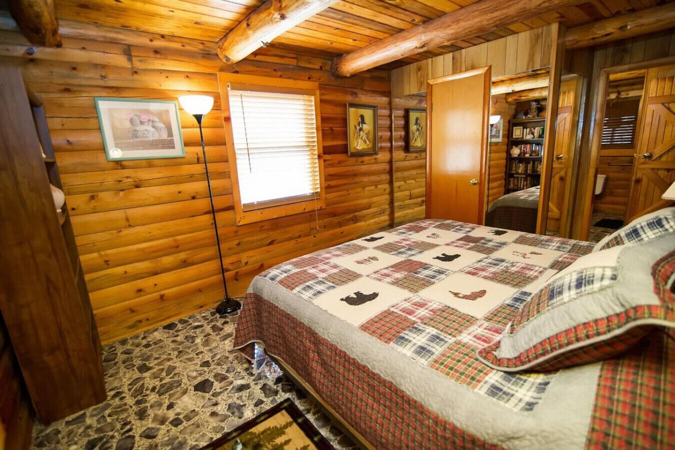 Une chambre dans une cabane en rondins, avec une couvre-lit matelassé, un sol en pierre, des murs en bois, une fenêtre avec des stores et une porte donnant sur un placard.