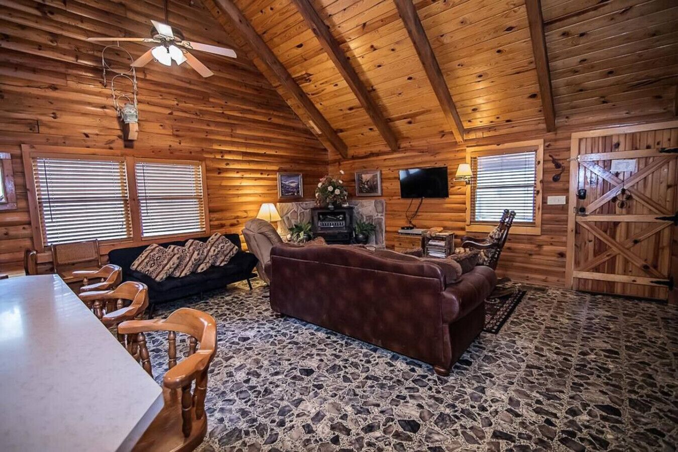 A log cabin interior with a stone-tiled floor, wooden furniture, a fireplace, and a television mounted on the wall.
