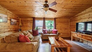 Een gezellig interieur van een chalet met een houten plafond, een plafondventilator, een televisie, een bank, een tweezitter en een houten salontafel.
