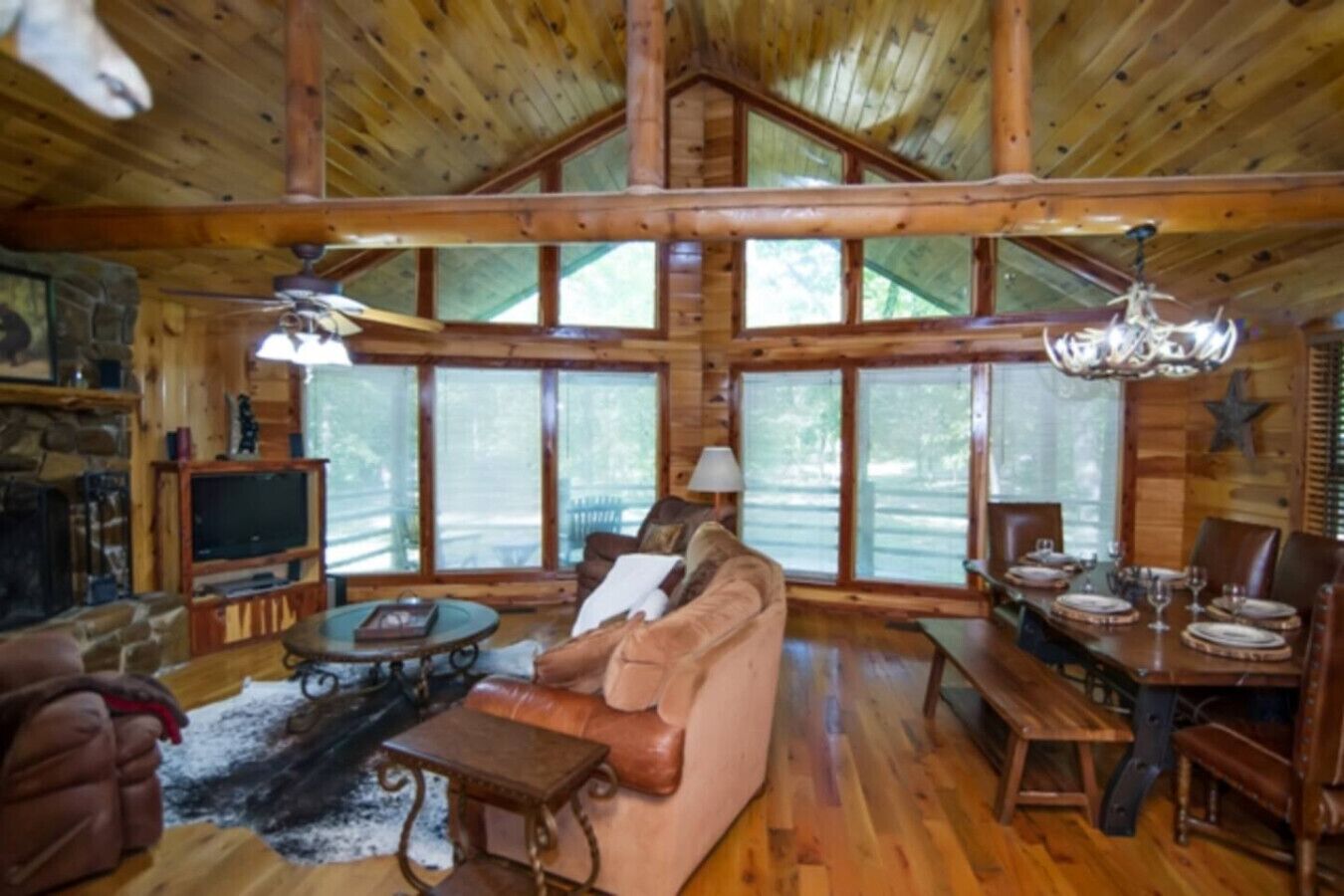 A log cabin interior with a wooden ceiling, stone fireplace, leather sofa, dining table set, and large windows.