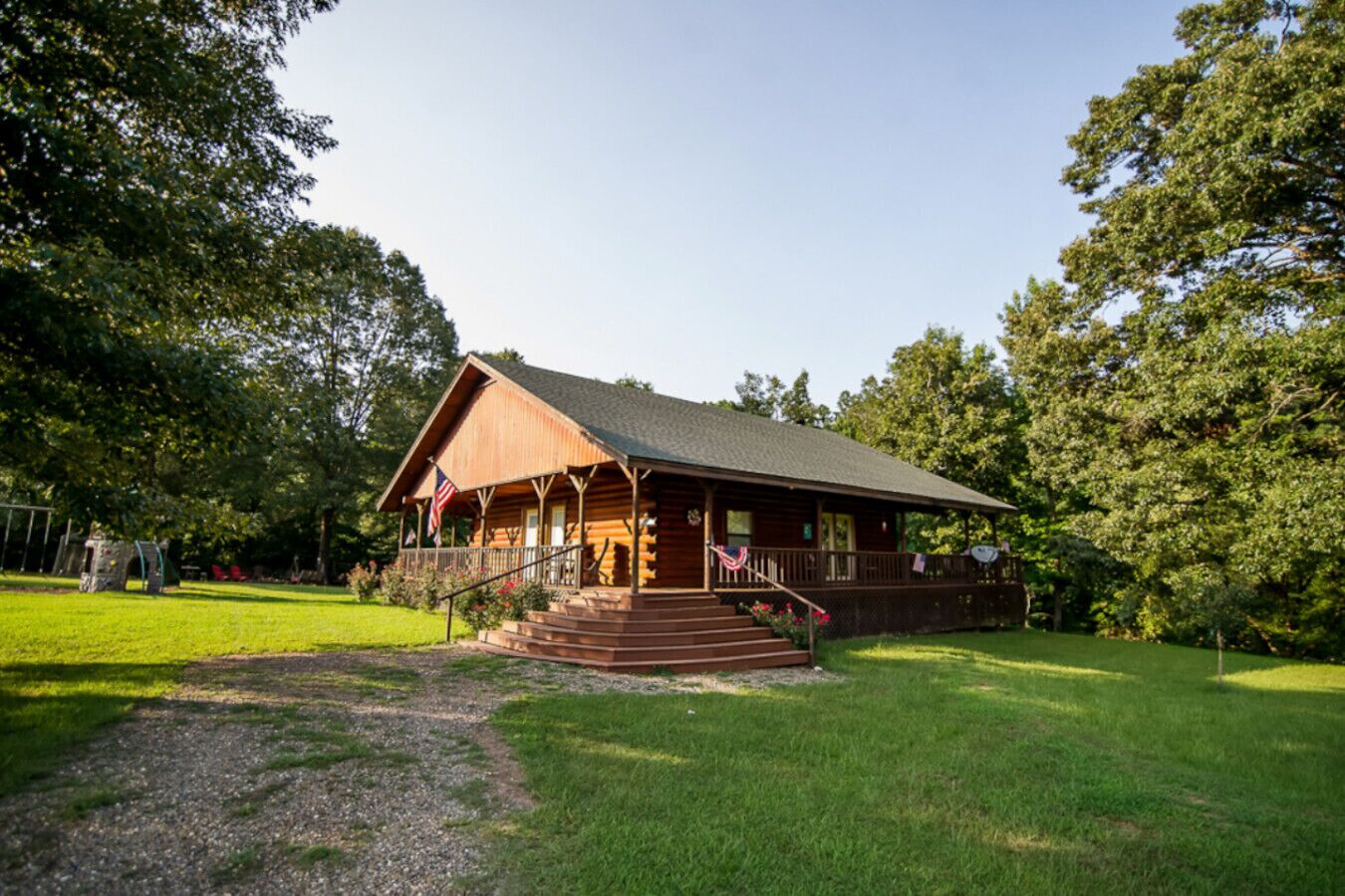 A wooden lodge with a porch and steps, surrounded by trees and a grassy area.