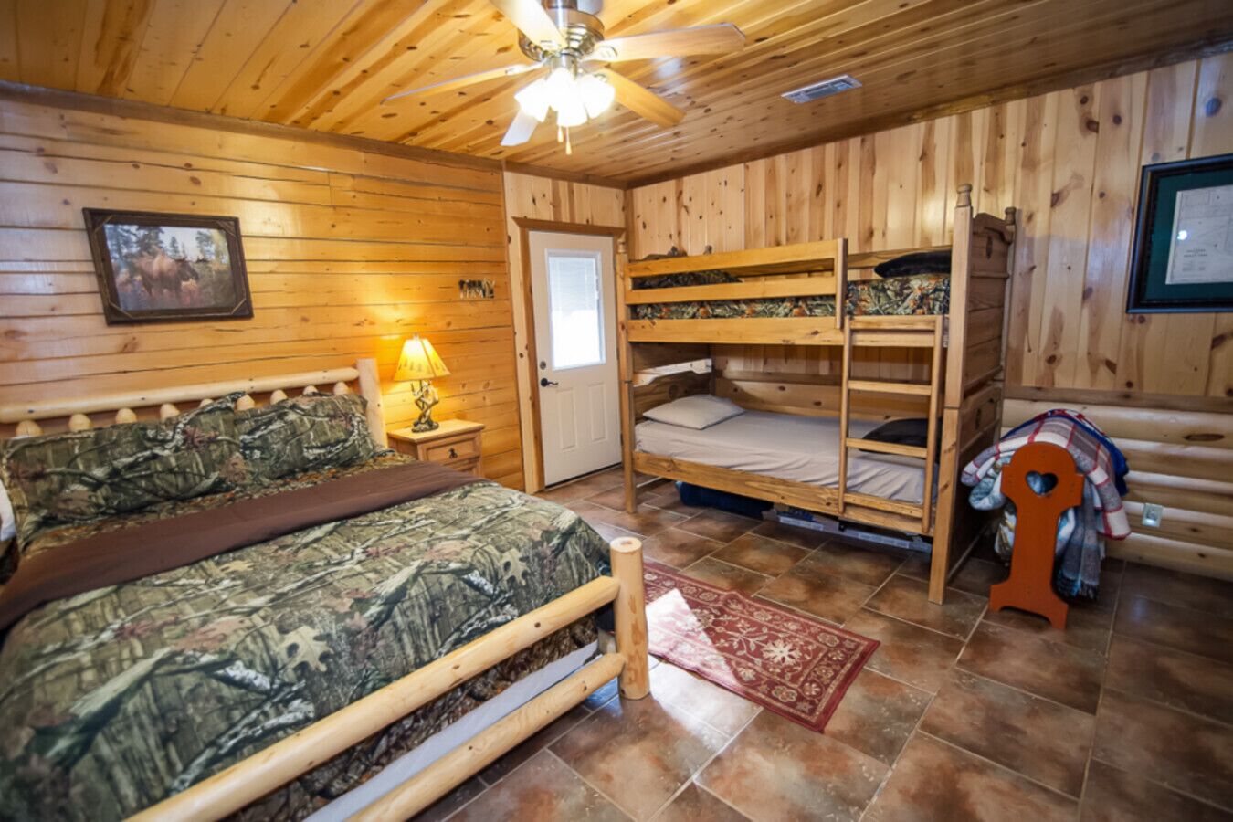 A cabin-style room with a bunk bed, a single bed with camouflage bedding, a wooden ceiling, and a red rug.