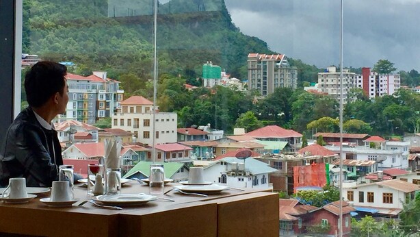 A dining area with a table set for two, overlooking a cityscape with buildings and a hill.