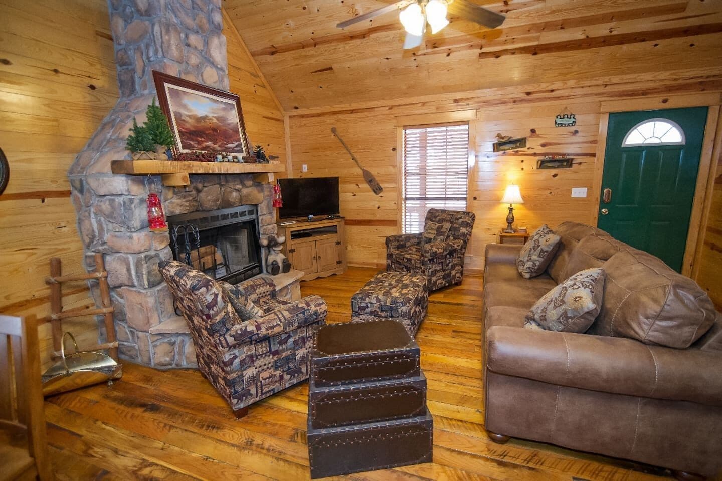 A cozy log cabin interior with a stone fireplace, a television, a green door, and a brown leather couch.