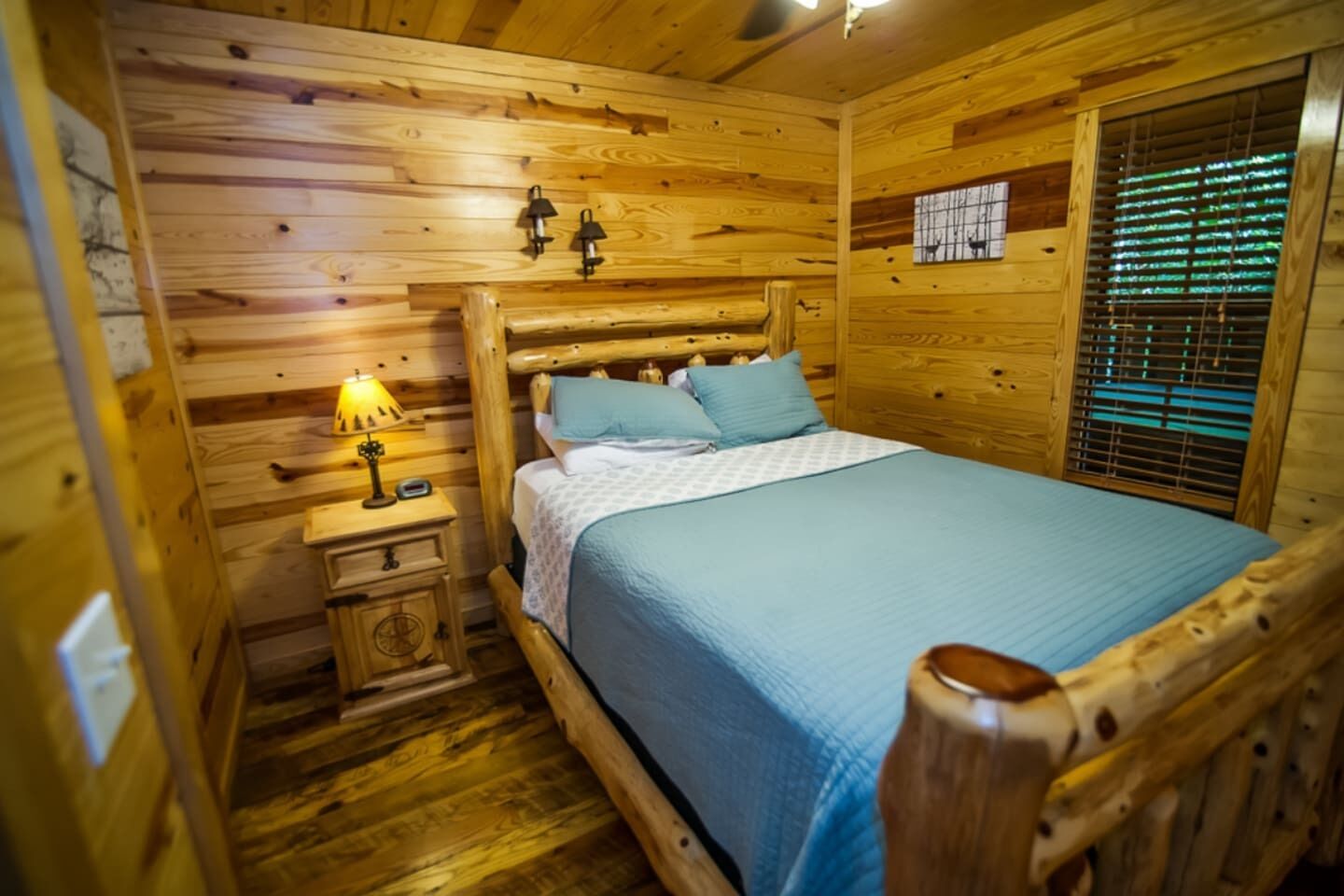 Habitació de cabana de fusta amb llit, tauleta de nit, làmpada i finestra amb persianes.