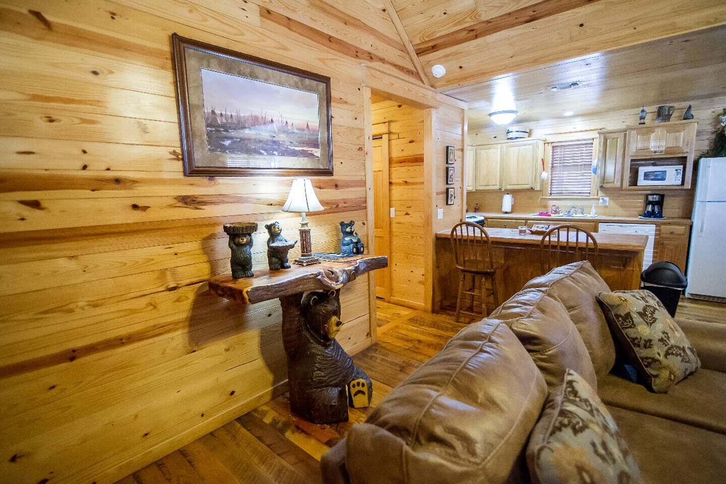 A cozy cabin interior with a wooden wall, a bear-themed side table, a leather couch, and a kitchen area with wooden cabinets and appliances.