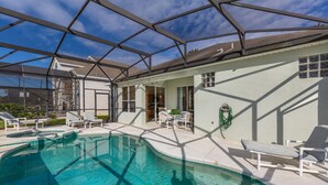 A swimming pool with lounge chairs, a covered patio area, and a view of a house in the background. - Enchanted Cottage by VillaDirect (Kissimmee)