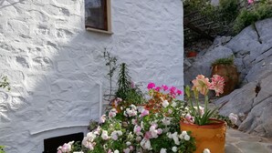 A white stone building with a window, surrounded by potted plants with pink and white flowers. - Arbeli House for idyllic sun & sea holidays in Mandraki Bay, Hydra (Idra)