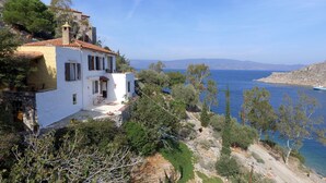 A white building with a red-tiled roof, a small patio with outdoor furniture, and a view of the sea and distant mountains. - Arbeli House for idyllic sun & sea holidays in Mandraki Bay, Hydra (Idra)