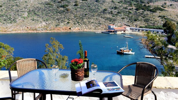 A table with a bottle of wine, a glass, and a magazine, overlooking a bay with boats and a building in the distance. - Arbeli House for idyllic sun & sea holidays in Mandraki Bay, Hydra (Idra)