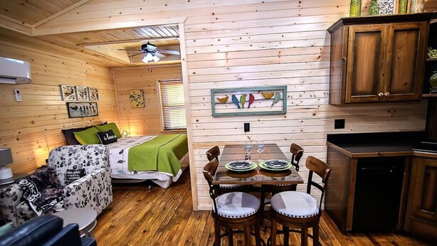 Interior acogedor de una cabaña con piso de madera, una mesa de comedor con sillas, un sofá, una cama con una manta verde y una zona de cocina con armarios de madera.