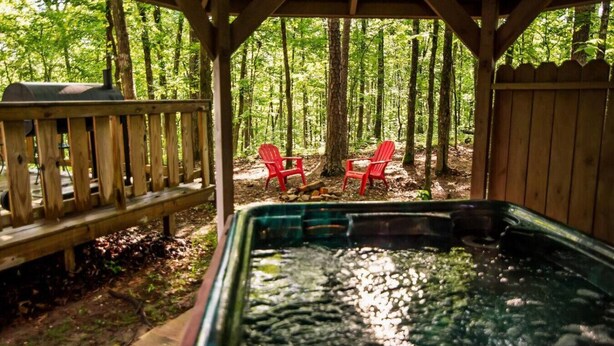 A hot tub in a wooded area with a wooden deck and two red chairs. - Papa Bear- Broken Bow/Hochatown- 1bd (Broken Bow)