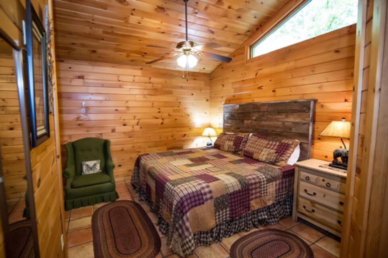 Un dormitorio de cabaña acogedor con techo de madera, un sillón verde, una cama con colcha, una mesita de noche con lámpara y una ventana.