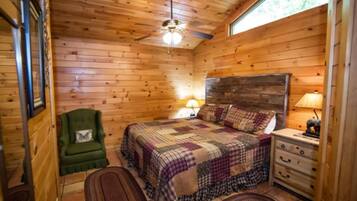 Un dormitorio de cabaña acogedor con techo de madera, un sillón verde, una cama con colcha, una mesita de noche con lámpara y una ventana.