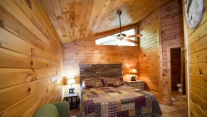 A cozy wooden cabin bedroom with a bed, a nightstand, a lamp, and a window. - Papa Bear- Broken Bow/Hochatown- 1bd (Broken Bow)