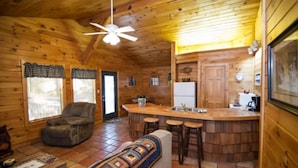A cozy cabin interior with a wooden ceiling, a bar area with stools, a sofa, a dining table, and a refrigerator. - Papa Bear- Broken Bow/Hochatown- 1bd (Broken Bow)