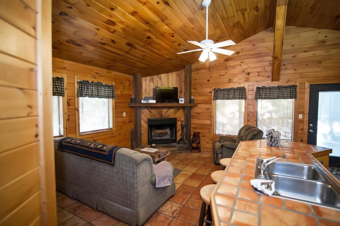 Interior acogedor de una cabaña con chimenea, un sofá, una mesa de comedor con fregadero y un ventilador de techo.