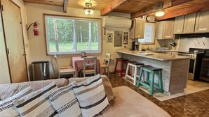 Acogedor interior de cabaña con sala de estar, cocina y un amplio ventanal con vistas a la vegetación.