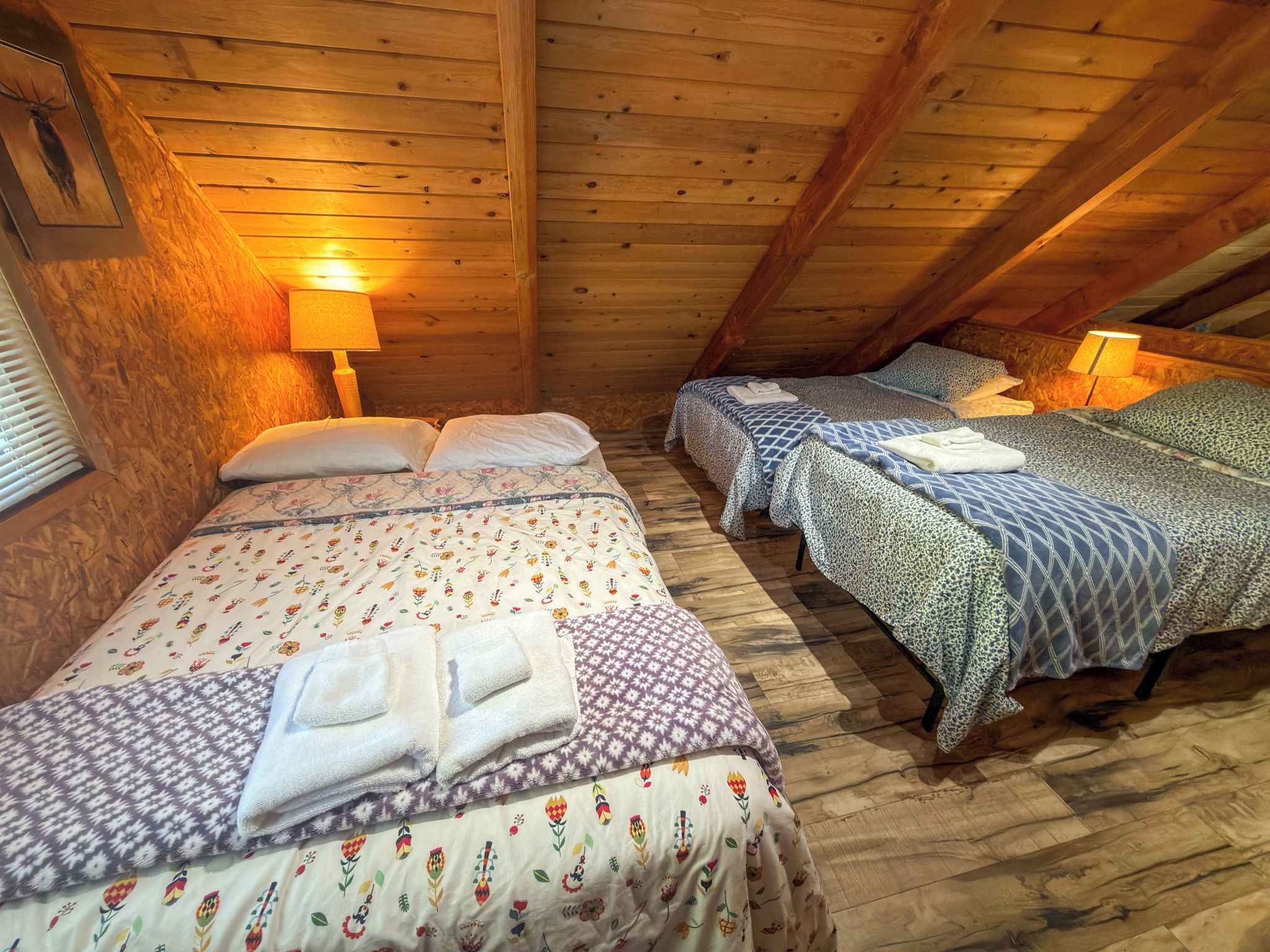 A cozy attic bedroom with wooden walls and sloped ceilings, featuring two beds with floral bedding and a lamp on each.