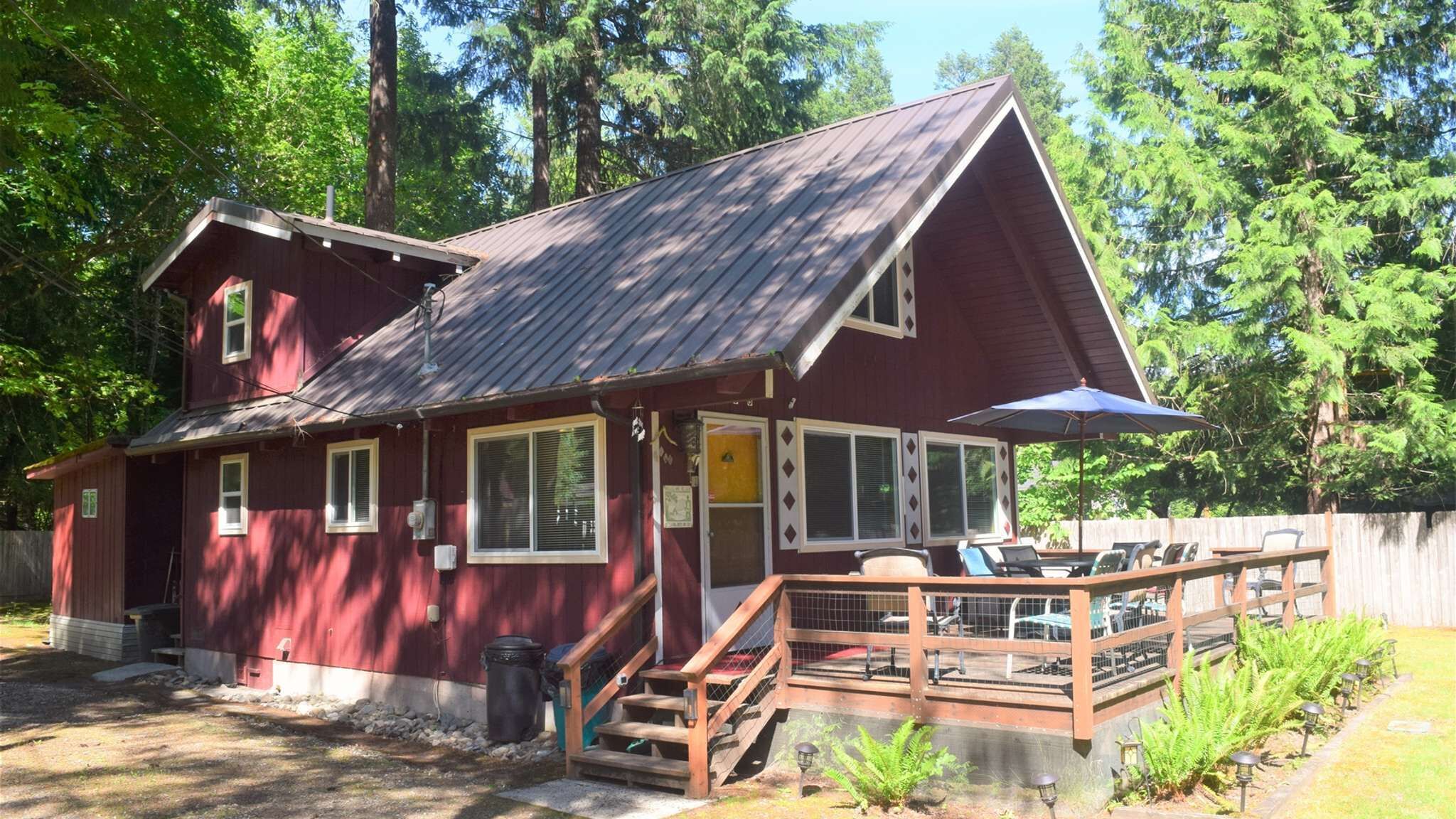 Eine zweistöckige rote Holzhütte mit überdachter Veranda, einer Eingangstür und einer kleinen Sitzgelegenheit im Freien.