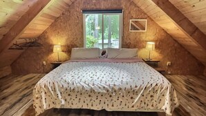 Un dormitorio acogedor en el ático, con techo inclinado, una cama grande con ropa de cama floral, dos lámparas de noche y una ventana que da a zonas verdes.
