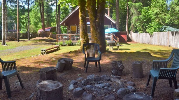 Un área de estar rústica al aire libre con una chimenea, rodeada de árboles y una terraza de madera.