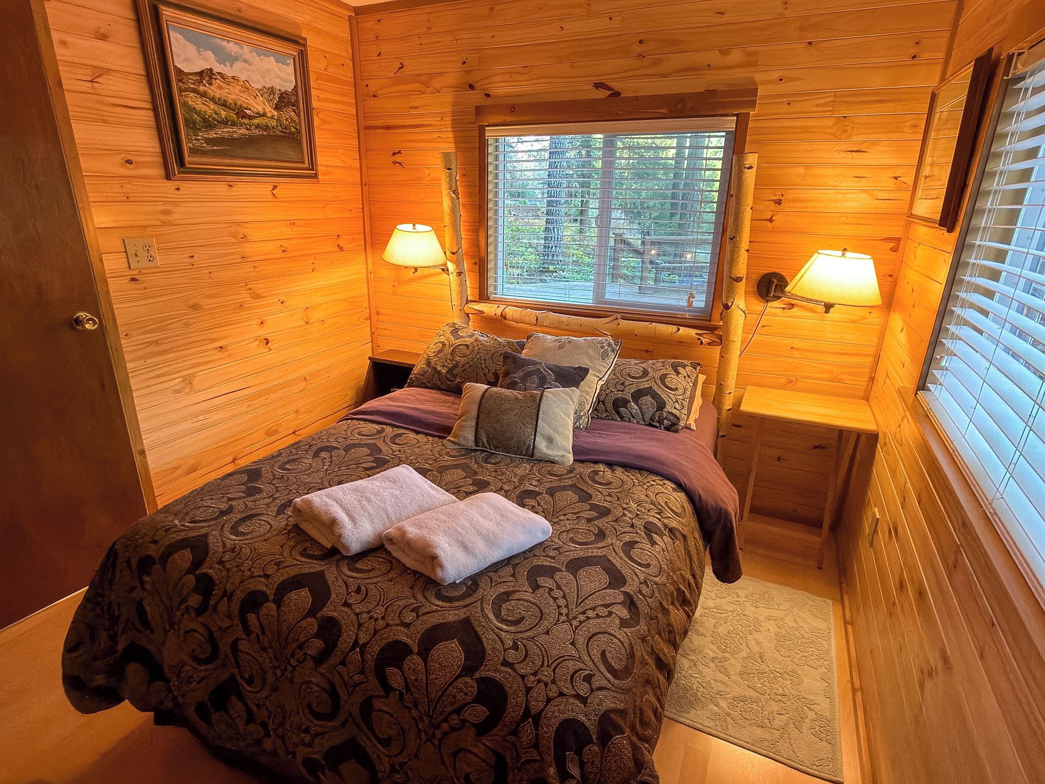 A wooden cabin bedroom with a bed, pillows, a window, and two wall-mounted lamps.