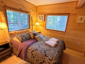 A wooden cabin bedroom with a bed, two windows, a lamp, and a framed picture on the wall. - Culps Cascade | Wi-Fi, Woodstove, Cedar Hot tub (Packwood)