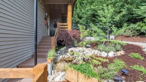 Ein Wohnhaus mit einer Holztreppe, die zu einer Veranda führt, einem Garten mit verschiedenen Pflanzen und einem Felsen, sowie einem Hintergrund aus immergrünen Bäumen.
