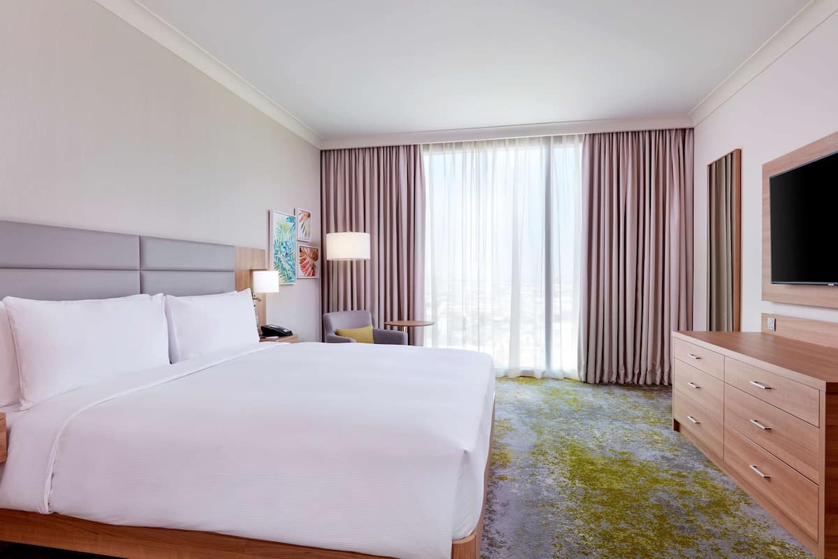 A hotel room with a large bed, a desk with a lamp, a TV, and a view of the city through the window.