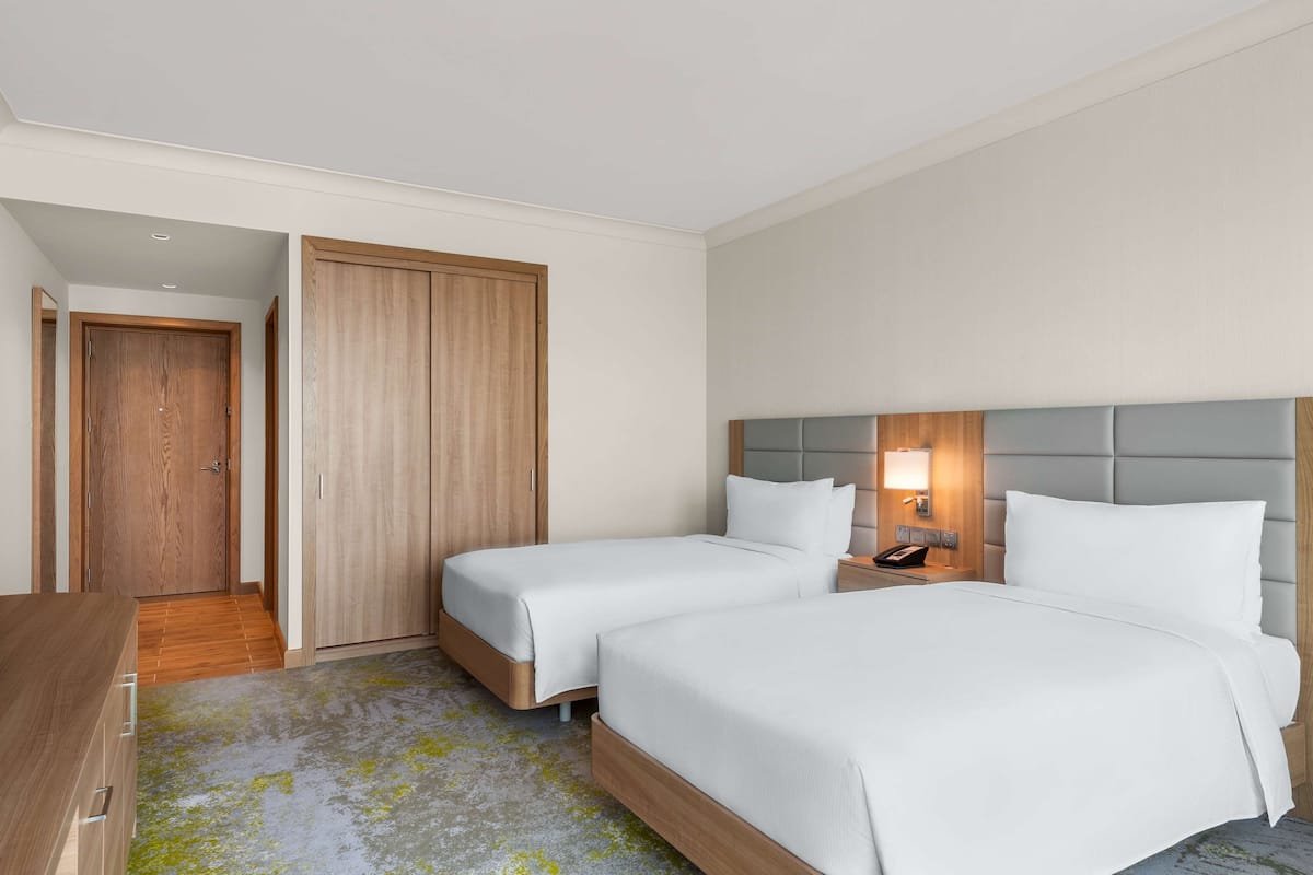 A hotel room with two beds, a wooden wardrobe, a desk, and a nightstand with a lamp.