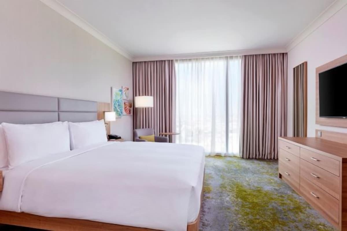 A hotel room with a large bed, a flat-screen TV, a desk with a lamp, and a view of the outdoors through the window.