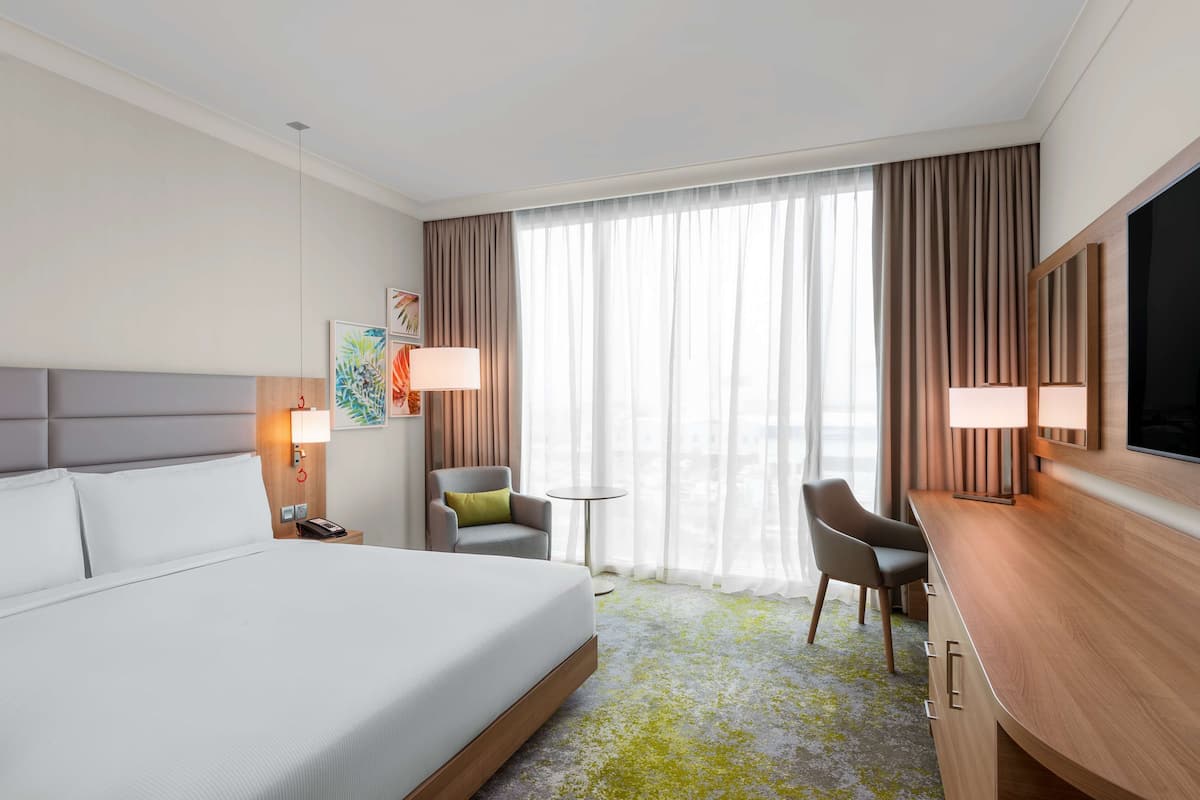 A hotel room with a large bed, a desk with a TV, a chair, a small table, and a window with curtains.
