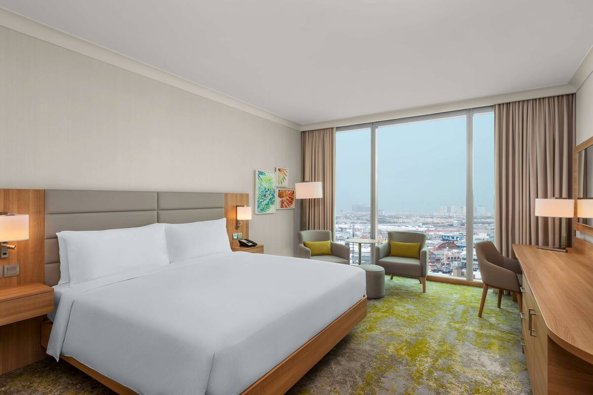 A hotel room with a large bed, a desk, a chair, a small table, and a view of the city.