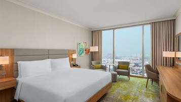 A hotel room with a large bed, a desk, a chair, a small table, and a view of the city.