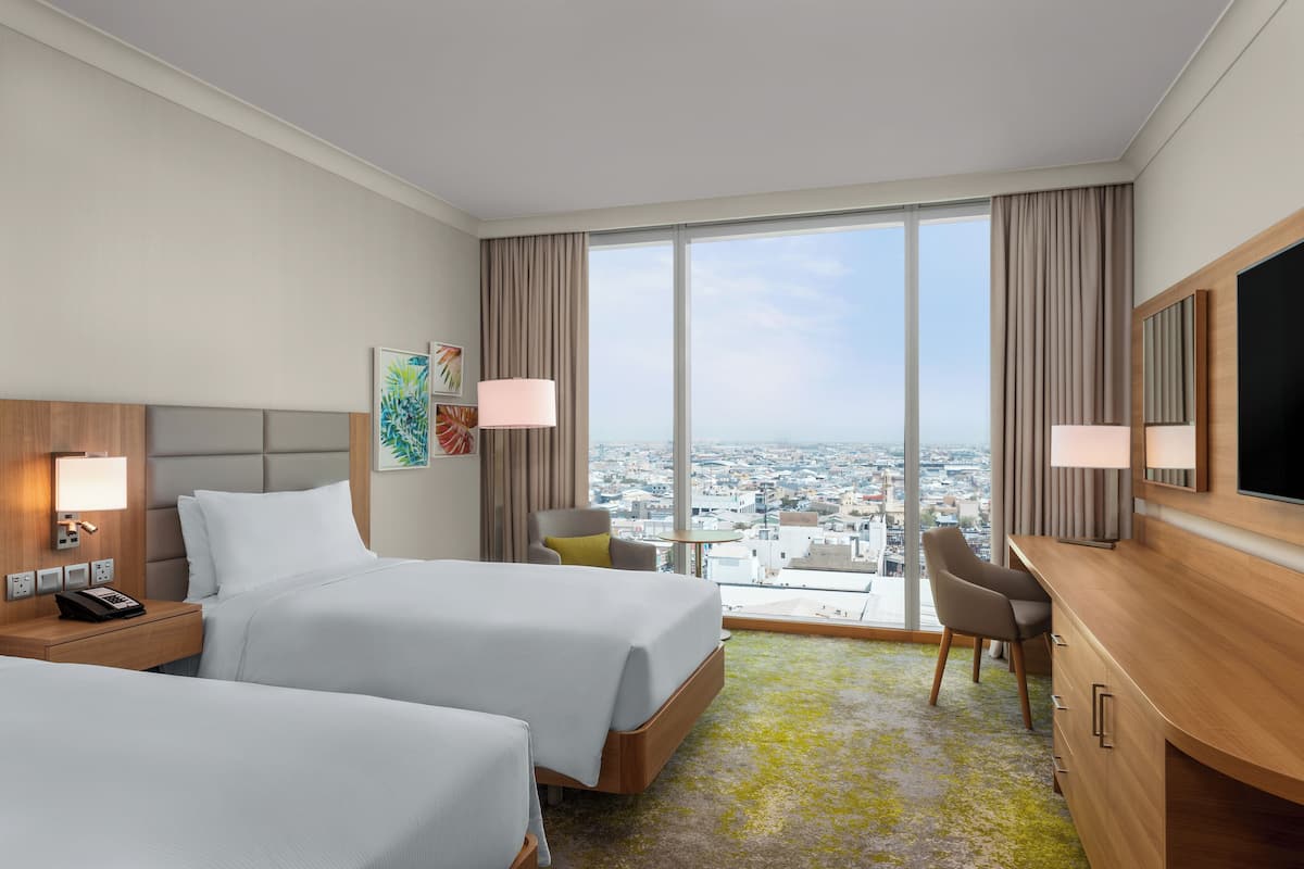 A hotel room with two beds, a desk, a chair, and a large window offering a city view.