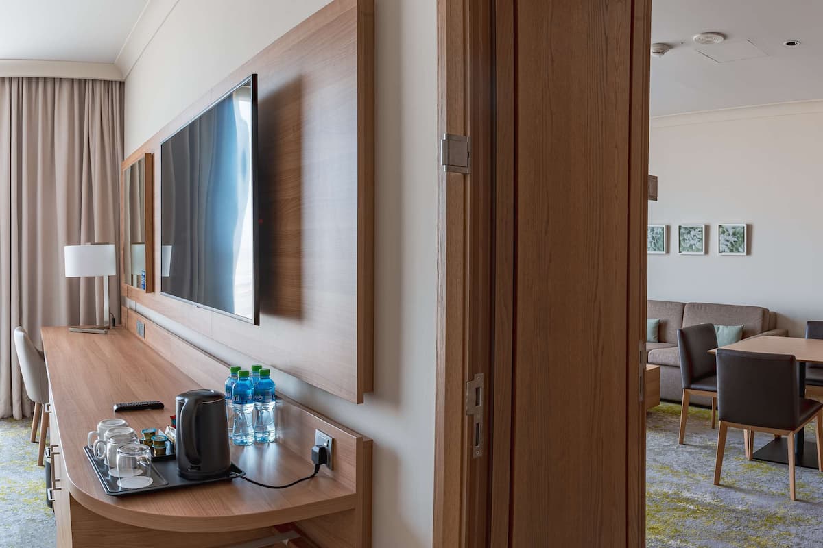 A hotel room with a wooden desk, a flat-screen TV, a sofa, a dining table with chairs, and a carpeted floor.