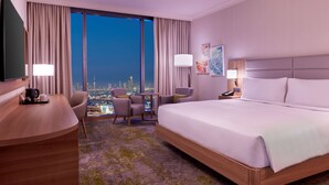 A hotel room with a large bed, a desk, a chair, and a view of a cityscape at night. - Hilton Garden Inn Kuwait (Kuwait City)