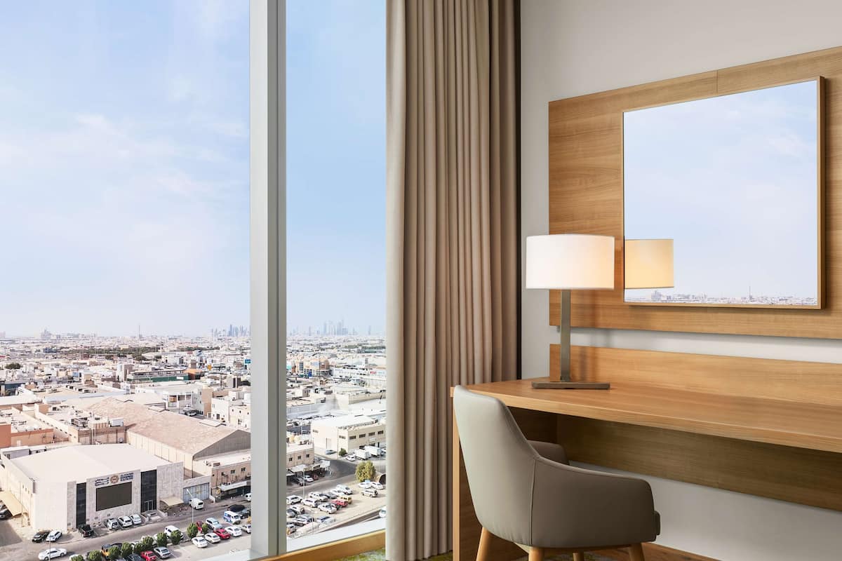 A hotel room with a large window offering a city view, a wooden desk with a lamp, and a mirror on the wall.
