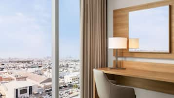 A hotel room with a large window offering a city view, a wooden desk with a lamp, and a mirror on the wall.
