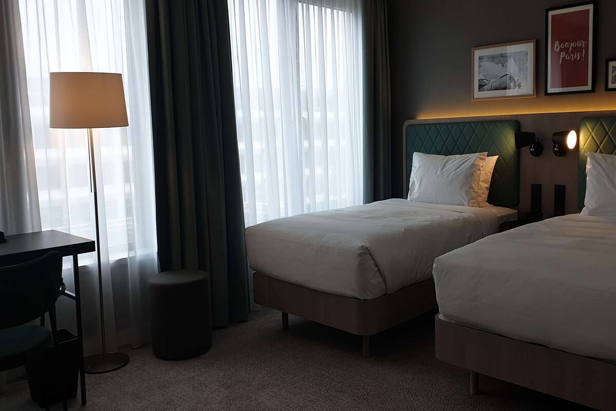 A hotel room with two beds, a desk, a lamp, and a view of the city.